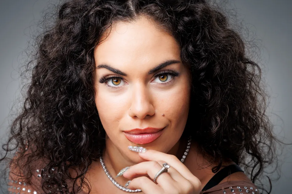 Close-up beauty portrait of a model with dark curly hair resting her hand under her chin against a grey studio backdrop