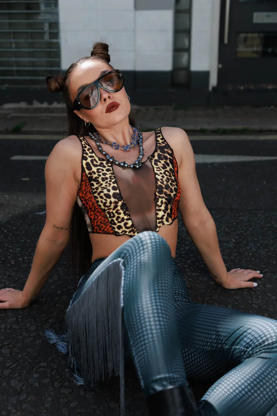 Street-style editorial portrait of a model in a leopard-print top and metallic trousers seated on a sunlit road
